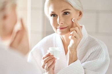 Eine Frau im Bademantel betrachtet sich lächelnd im Spiegel, während sie Creme aufträgt.