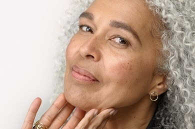 Reife Frau mit grauen Locken berührt sanft ihr Gesicht und zeigt gepflegte Haut.