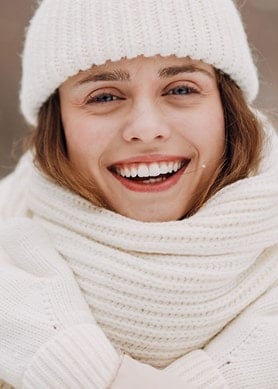 Eine lächelnde Frau mit weißer Mütze, weißem Schal und weißen Handschuhen in einer winterlichen Landschaft