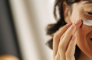 Eine lächelnde Frau trägt sich weiße Creme auf die Wange auf