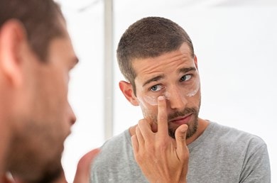 Mann mit kurzen Haaren steht vor dem Spiegel und trägt sich mit dem Finger Augencreme auf