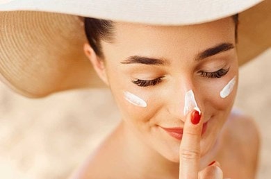 Eine junge Frau mit Sonnenhut, die sich jeweils einen Streifen Sonnencreme auf beide Wangen und die Nase aufträgt